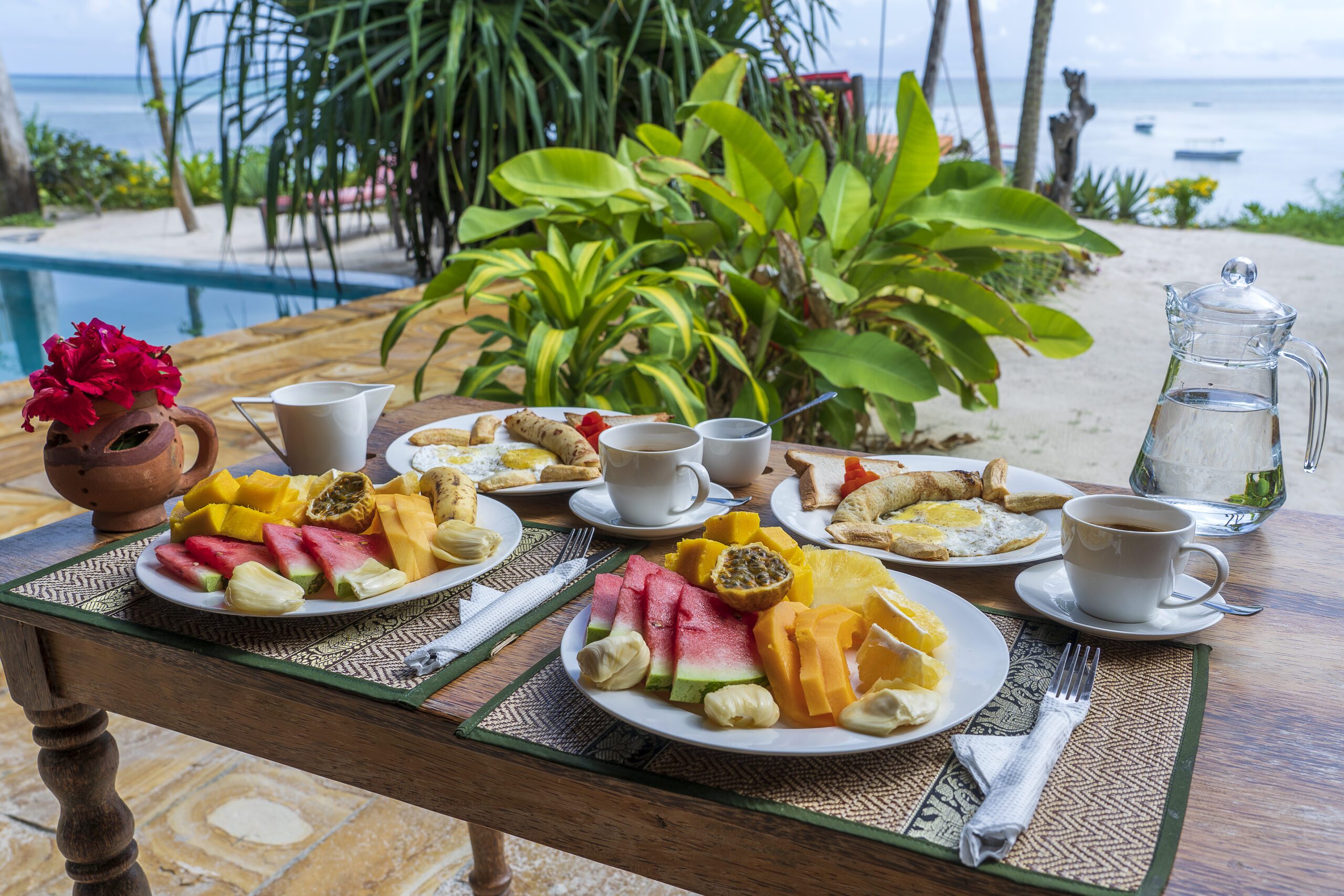 Tropical breakfast of fruit, coffee and scrambled eggs and banana pancake for two on the beach near sea. Top view, table setting.